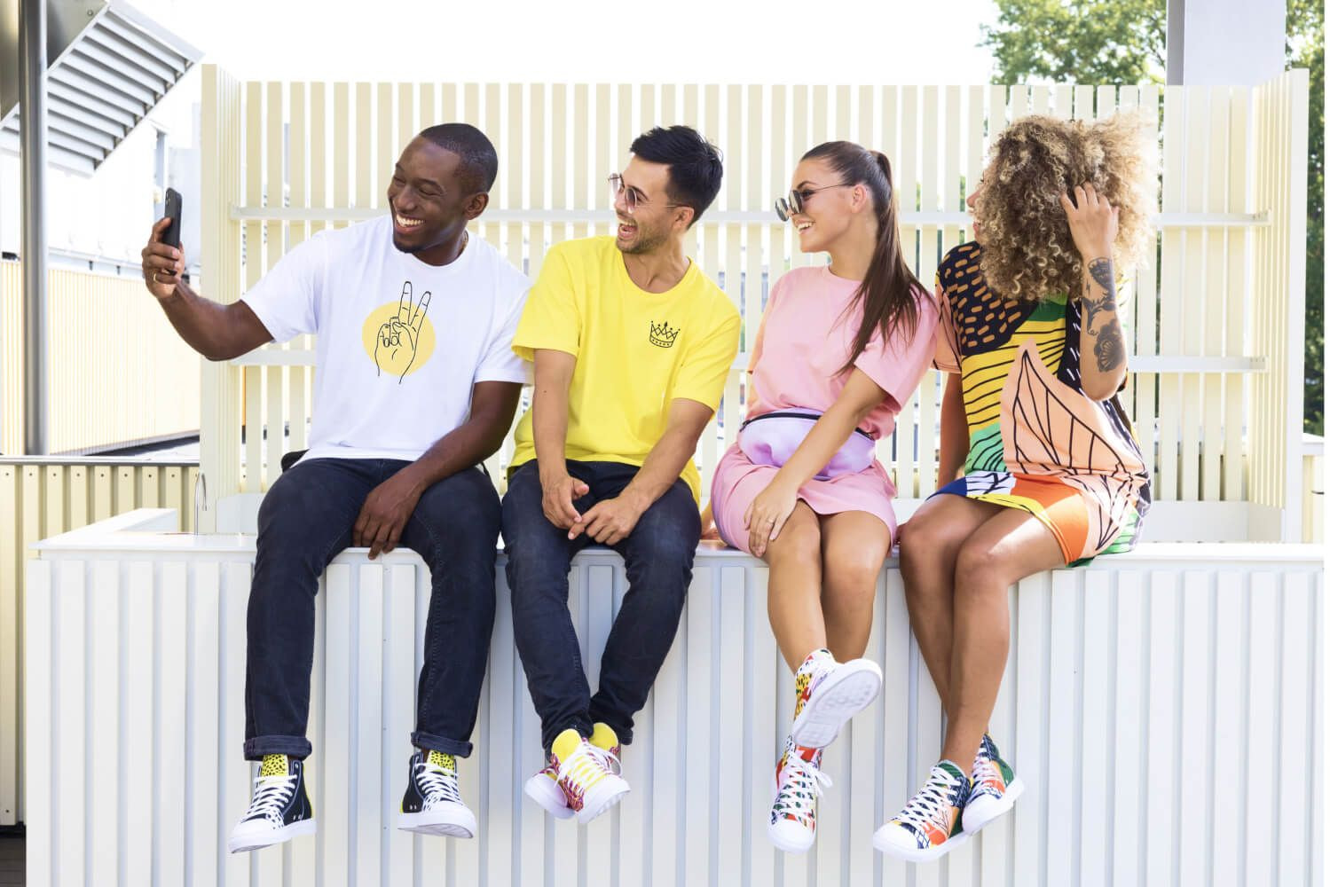 Amis étudiants portant des tenues de style urbain colorées prenant des photos avec leur smartphone et portant des lunettes de soleil.