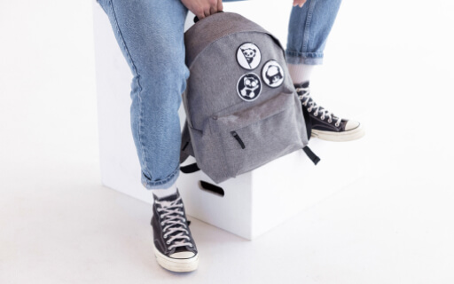 Model in jeans and black high sneakers shoes sitting on the white cube holding a grey backpack with black-and-white patches.
