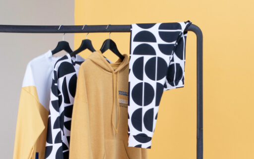 Percha con sudaderas negras, blancas y amarillas y leggins sobre un fondo de estudio gris y amarillo.